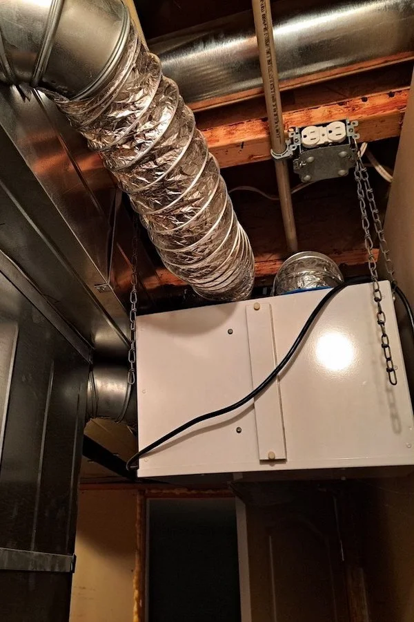 Un échangeur d'air (VRC) blanc installé sur le mur du sous-sol, avec des conduits de ventilation argentés attachés.