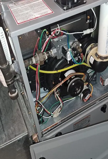 Une vue rapprochée de l'intérieur d'une fournaise au gaz résidentielle à haute efficacité avec le panneau avant retiré. On y voit le moteur de tirage, l'ensemble de la valve à gaz, ainsi que divers câblages électriques et capteurs.