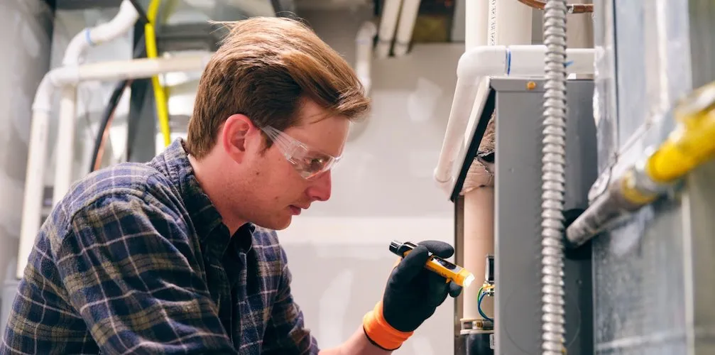 Un technicien portant des lunettes de sécurité et des gants inspecte la fournaise. Il utilise une petite lampe de poche pour examiner les composants internes et les connexions électriques.