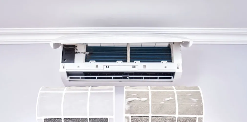 Une vue d'ensemble de l'unité intérieure de climatisation avec son couvercle relevé et les filtres retirés. En dessous, les deux filtres détachés sont visibles, montrant une accumulation importante de saleté et de débris.