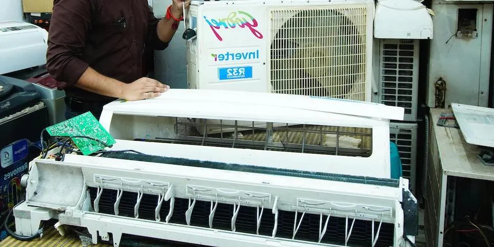 Un technicien dans un atelier travaillant sur un climatiseur démonté. Les composants internes, y compris la carte de circuit imprimé et le ventilateur, sont exposés pour un nettoyage en profondeur ou une réparation.