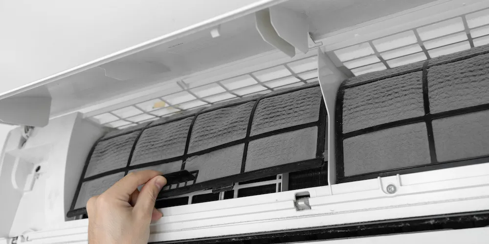 Gros plan d'une main retirant un filtre à air empoussiéré d'un climatiseur mural blanc pour le nettoyage.