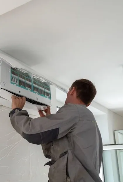Un technicien en tenue de travail grise effectue l'entretien ou l'installation d'un climatiseur mural. Le panneau avant est ouvert, laissant apparaître les filtres et les composants internes.