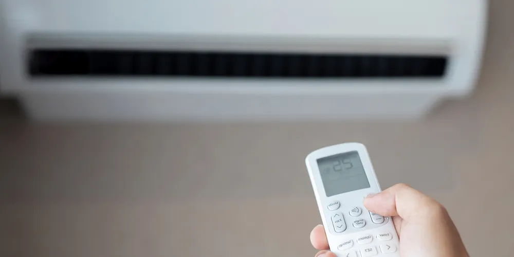 Un gros plan d'une main tenant une télécommande dirigée vers un climatiseur. L'écran de la télécommande affiche un réglage de température de 25°C.