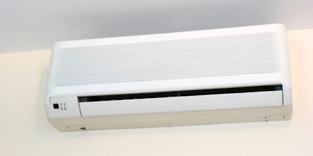 Une vue de face d'un climatiseur mural compact blanc installé en hauteur. L'appareil possède une grille supérieure texturée pour l'entrée d'air et une fente inférieure pour la diffusion du froid.