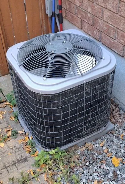 Une vue en plongée d'une unité de condensation de climatisation extérieure grise et carrée, posée sur un patio de gravier et de pierre à côté d'un mur en briques. Quelques mauvaises herbes et feuilles mortes sont éparpillées à la base.