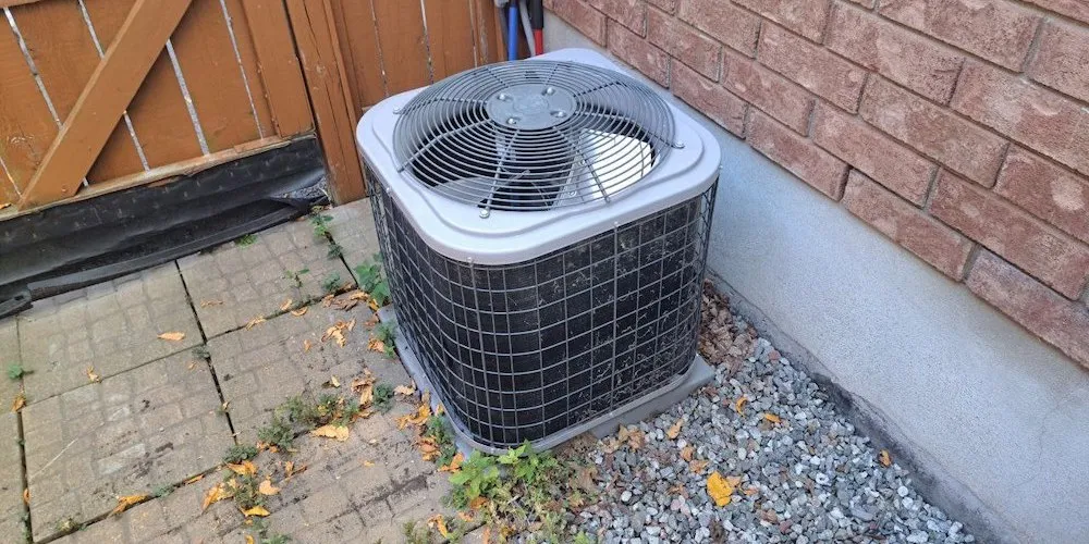 Un climatiseur central extérieur gris et carré, posé sur un sol en gravier à côté d'une maison en briques. Un portail en bois et des pavés en pierre sont visibles en arrière-plan.