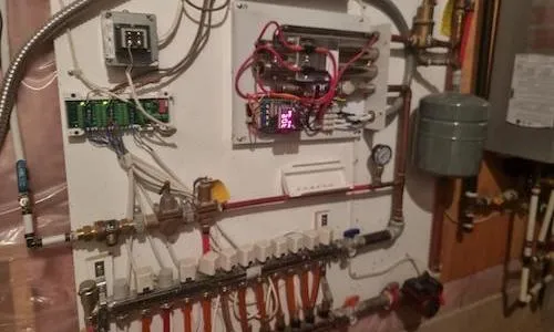 Un système de contrôle de plancher chauffant hydronique mural. On y voit un collecteur avec plusieurs tubes PEX oranges, des vannes de zone blanches, une pompe de circulation, un réservoir d'expansion et un régulateur de température numérique avec un câblage rouge.