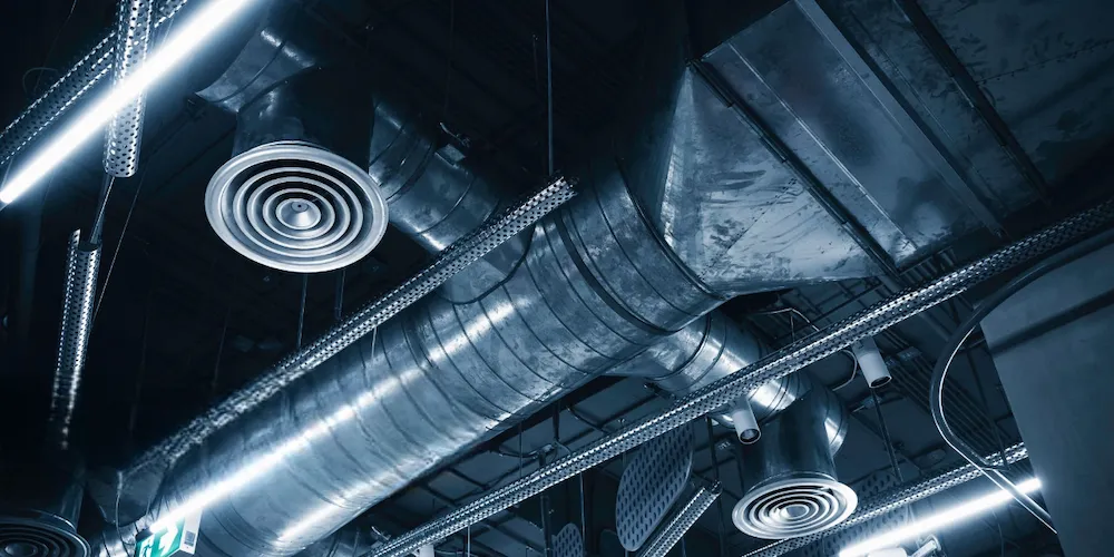 Vue en contre-plongée d'un plafond industriel montrant des conduits de ventilation en métal argenté, des diffuseurs d'air circulaires et un éclairage LED linéaire brillant.