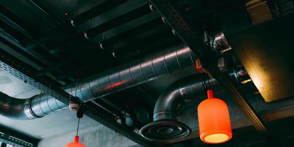 Intérieur sombre avec infrastructures apparentes, incluant des conduits de ventilation cylindriques, des conduits électriques et deux suspensions lumineuses orange.