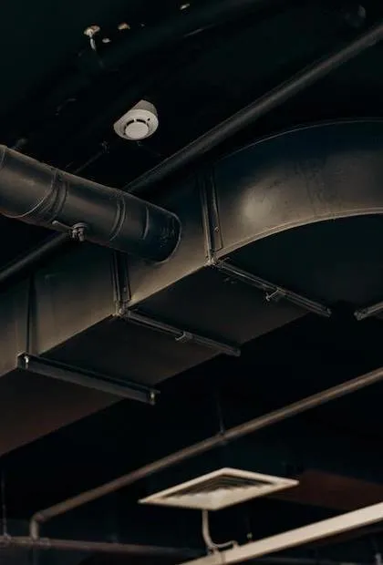 Vue de conduits de climatisation circulaires et rectangulaires peints en noir sur un plafond sombre, avec un détecteur de fumée blanc et une grille d'aération à ailettes en arrière-plan.