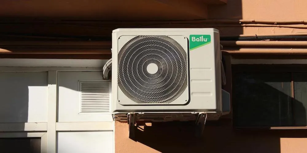 Un plan moyen d'un compresseur de climatisation extérieur de marque Ballu, de couleur blanche, fixé à un mur extérieur beige à l'aide de supports métalliques. L'unité présente une grande grille de ventilateur circulaire et se trouve à côté d'une fenêtre et d'une porte blanche munie d'une petite grille d'aération carrée.